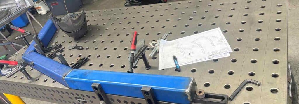 Close-up of a Demmeler welding table's modular grid surface with clamped fixture and manufacturing drawing at Fabworx