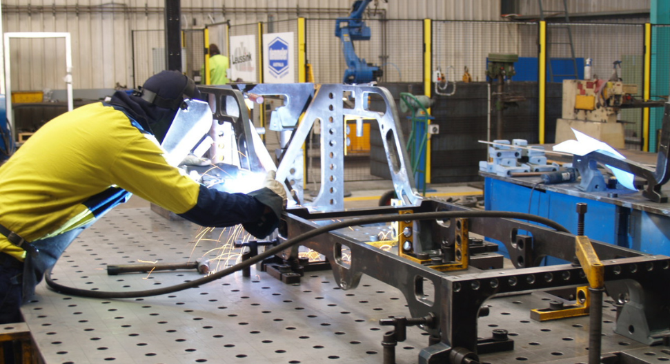Welder in hi-vis jacket and welding helmet working on a steel fabrication frame on a Demmeler welding table, with welding sparks and a robotic welding arm visible in the background