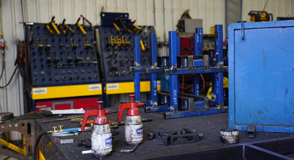 Fabrication workshop workbench with welding tools and spray bottles in the foreground, Demmeler fixturing boards and blue steel frame racking in the background
