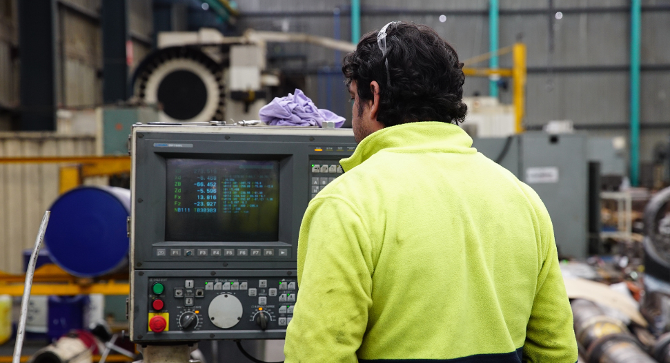 Machinist in hi-vis jacket viewed from behind, programming a CNC control panel with G-code display, heavy machinery in background