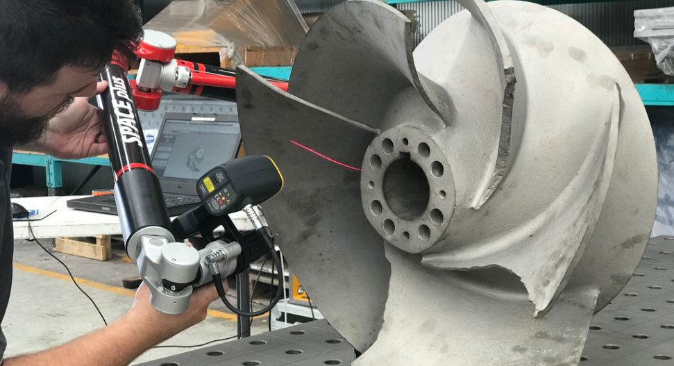 Technician using a Space Plus portable CMM arm with laser scanner to inspect a large marine propeller, with CAD software visible on a laptop in the background