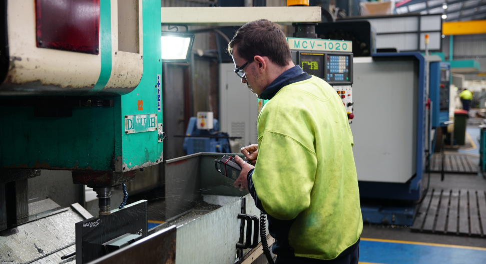 Machinist in hi-vis jacket operating a CNC vertical milling machine with a handheld pendant controller, with an MCV-1020 CNC machining centre visible in the background