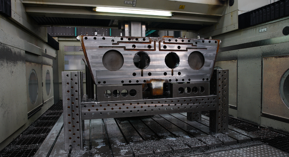 Large machined steel housing or gearbox casing fixtured on a modular tombstone setup inside a CNC horizontal boring mill, with coolant residue visible on the workpiece and machine bed