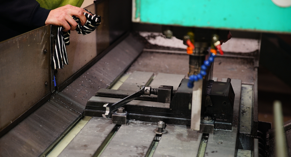 Machinist wiping down a milling machine table with a workpiece clamped in a vice, coolant nozzles visible in the background