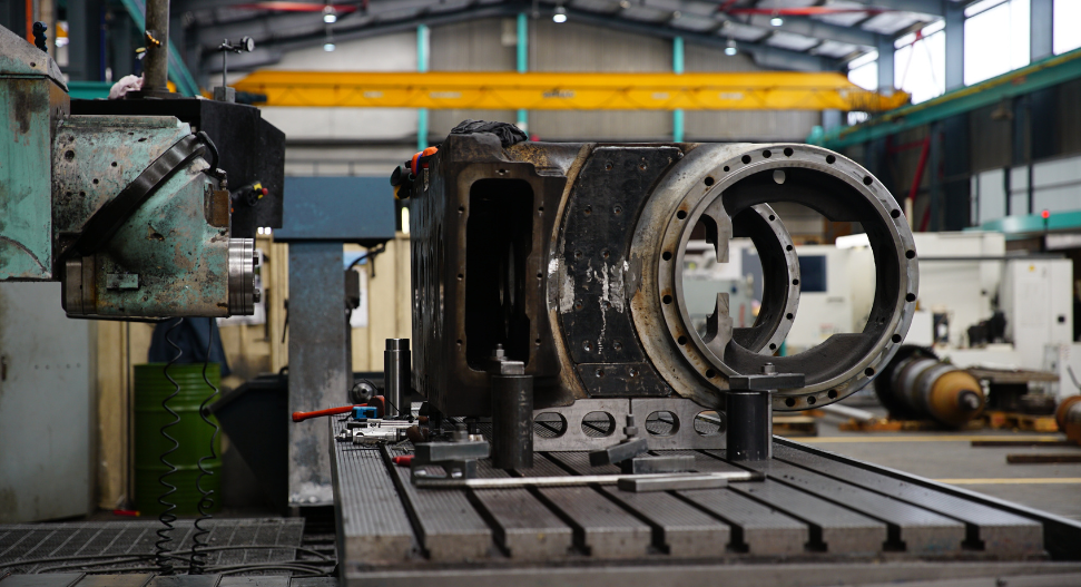 Large heavy steel gearbox or industrial housing casing set up on a horizontal boring mill table for precision machining, in Leussink engineering workshop with overhead crane visible