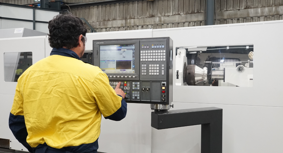 CNC machinist operating an Okuma CNC lathe control panel in a heavy industry workshop