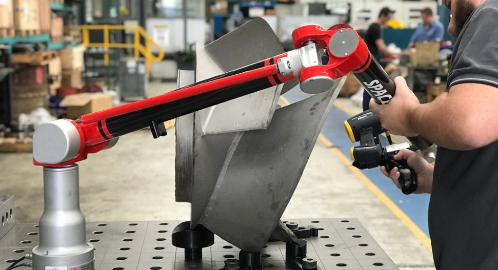A technician using a portable red articulated coordinate measuring arm to 3D scan a metal component on a fixture table in a manufacturing facility.