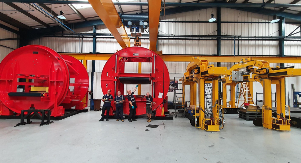 Four engineers in high-visibility vests standing beside large red circular industrial reels and yellow handling equipment inside a manufacturing warehouse