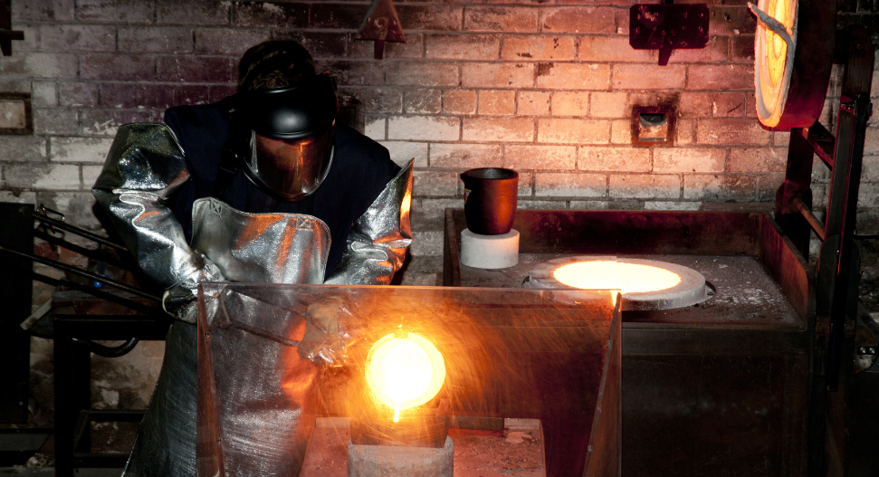 A foundry worker in full heat-protective gear and face shield pouring