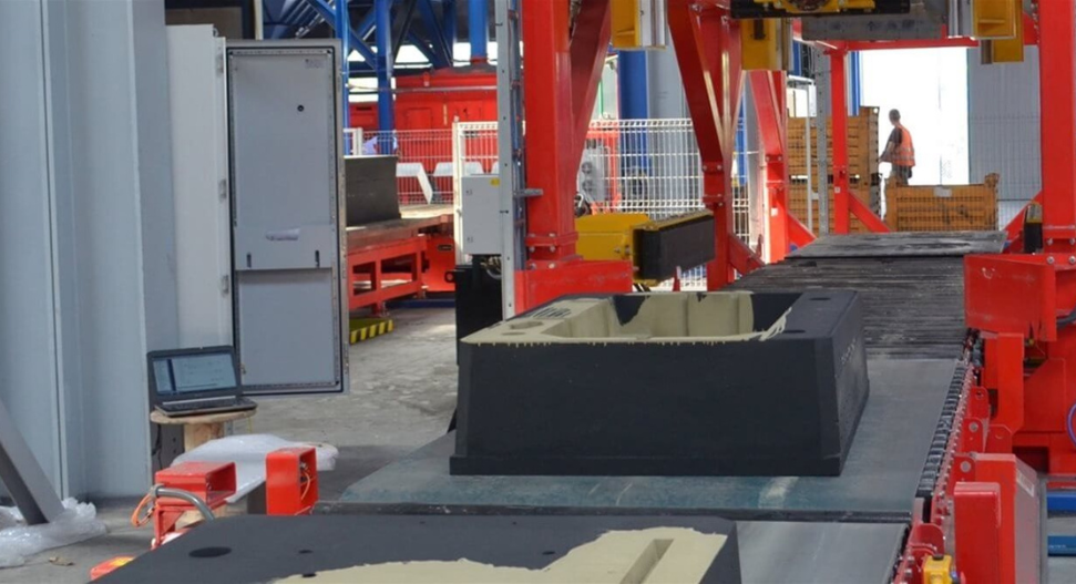 Industrial manufacturing facility with red gantry crane system handling large black moulded components on a production floor, with a worker visible in the background.