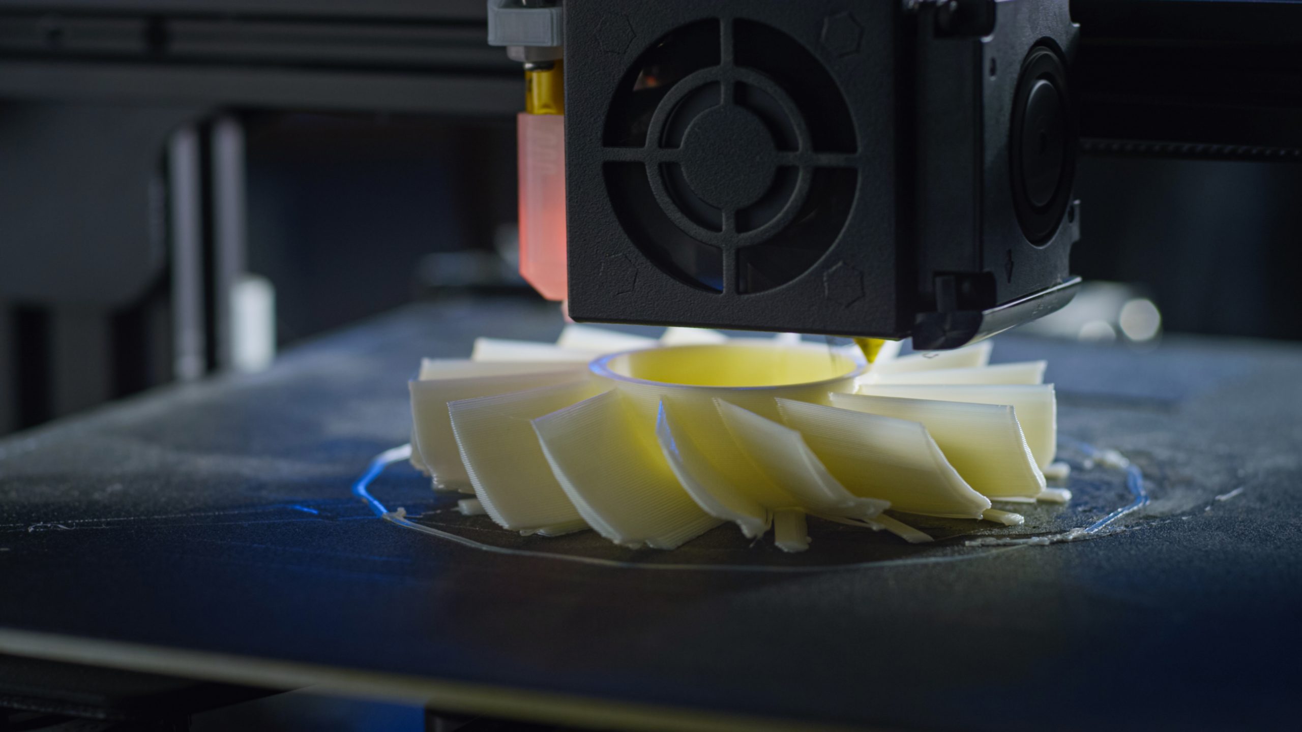 Close-up of a 3D printer extruder head printing a yellow spiral turbine or impeller component on a build plate
