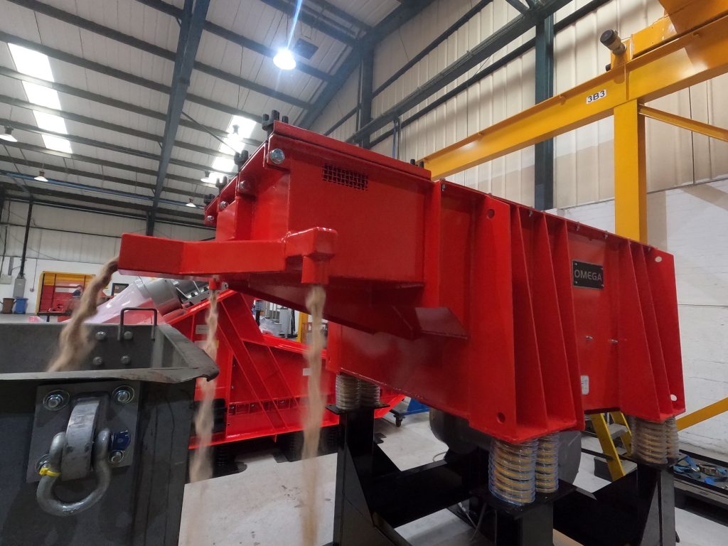 Omega Sinto vibratory shakeout sand reclamation system discharging reclaimed sand from a red industrial separation unit inside a modern foundry facility.