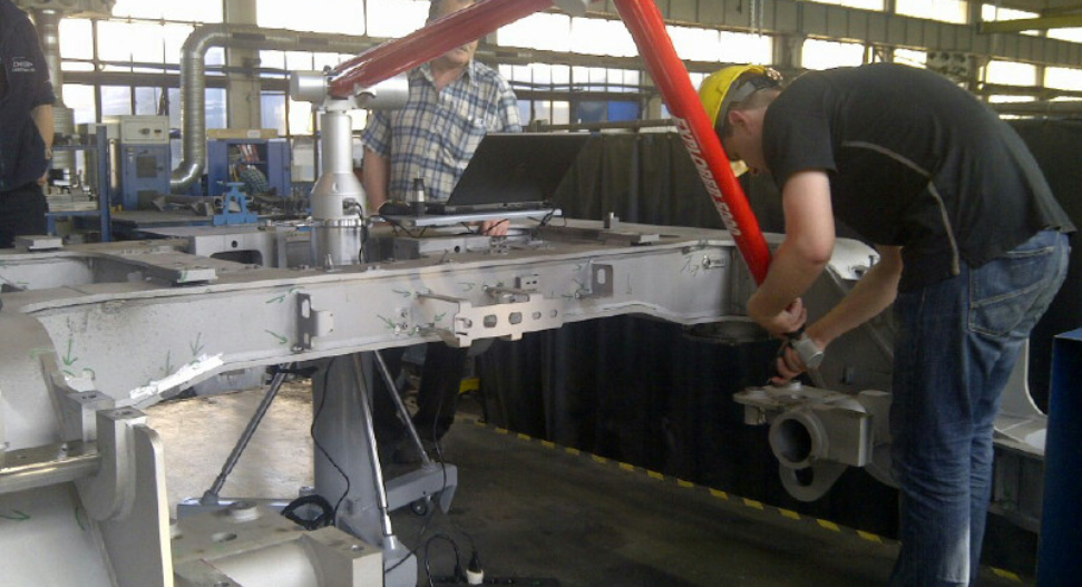 Technicians using an articulated arm CMM to inspect a large metal vehicle chassis frame in a workshop